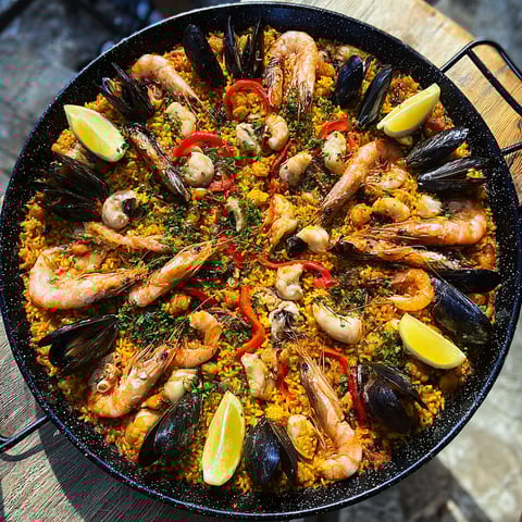 Une immense paella pleine de fruits de mer, avec tomates, poivrons, langoustines et oignons, posée dehors sur une grande planche en bois.